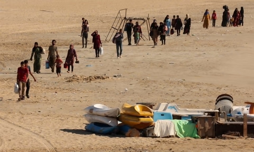 A line of displaced people carrying scant possessions walk along a beach. The photo was taken near Tyre, Lebanon. &10;&10;Image credit: Aziz Taher for Reuters