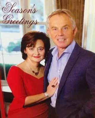 A grimacing couple poses together indoors, with the woman in a red dress and the man in a blue suit, accompanied by the text Season's Greetings it’s Tony and Cherie Blair.