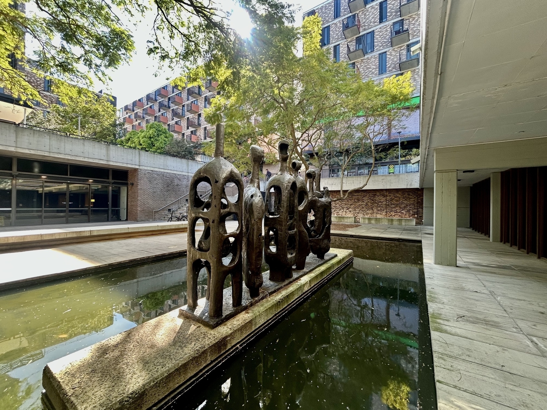 A group of abstract bronze sculptures in expressionist style stands on a rectangular platform surrounded by a shallow pond in a building courtyard. A jacaranda’s branches are visible, partly shading the area.