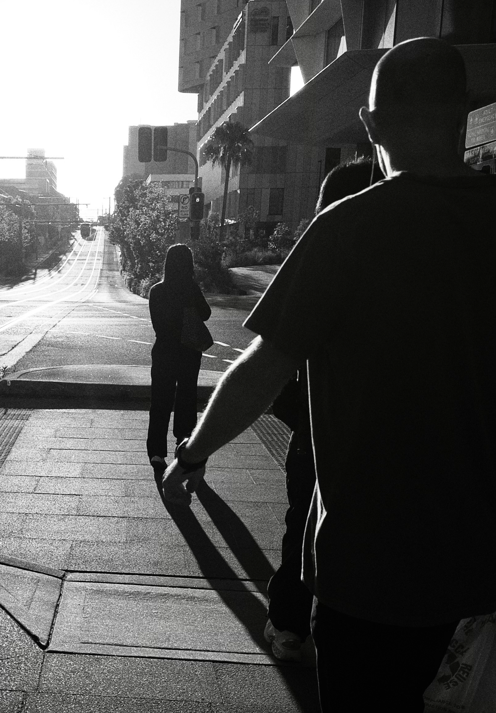 A person walks on the footpath facing away while another figure walks nearby on a sunlit street. A light rail line stretches off into the distance on the left of the photo. 