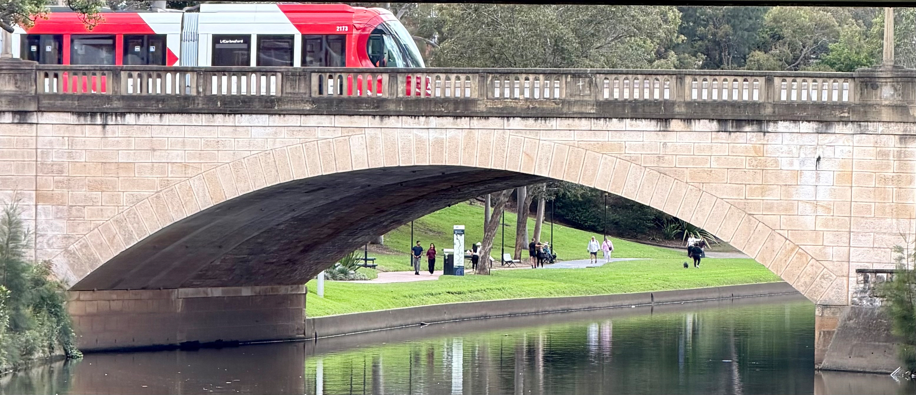 A tram is passing over a stone arch bridge with people walking and relaxing in a park area along a calm river. It’s the Parramatta light rail over the Parramatta River. 